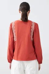 Remera blanca de manga larga con detalles bordados en tonos azules y verdes en el pecho y hombros. Presenta un escote en V con cordones ajustables terminados en borlas y puños elásticos.