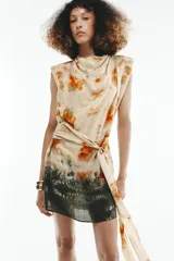 Vestido corto sin mangas de cuello redondo, con estampado tie-dye en tonos beige, naranja y verde oscuro. Presenta un lazo para atar en la cintura y cierre trasero con cremallera oculta.