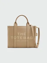 Cartera tipo tote de cuero granulado color tostado, con diseño estructurado y rectangular. Presenta doble asa superior fija y una correa larga ajustable y extraíble. En el frente, destaca el logo estampado con la inscripción 'THE TOTE BAG MARC JACOBS'.