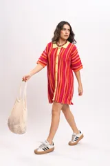 Vestido camisola corto de tejido fresco de algodón, con diseño a rayas verticales en tonos rojos, naranjas y fucsias. Cuenta con cuello polo y botonadura frontal en tono amarillo claro, y mangas cortas holgadas.