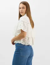 Camisa de algodón blanca con cuello clásico y cierre frontal de botones. Presenta un diseño con capas superpuestas y volados, combinando paneles de tela lisa con tejido bordado suizo de motivos calados y una cenefa decorativa. Posee mangas cortas y una silueta acampanada.