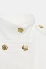 Chaqueta corta blanca de estilo militar, con cuello subido, manga larga y trabillas desflecadas en los hombros. Cuenta con cierre frontal cruzado decorado con botones metálicos dorados.