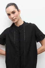 Vestido corto negro de corte amplio, con cuello mao, mangas cortas con volados y detalles de vainillas verticales en el frente. Cuenta con un volante fruncido en el ruedo.