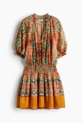 Vestido túnica corto y oversized de tejido plano con estampado tipo paisley y floral en tonos naranjas y azules. Presenta cuello redondo fruncido con abertura en V y lazos finos al frente, mangas cortas abullonadas con puños elásticos, cintura fruncida y falda con vuelo.