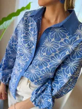 Camisa de manga larga con diseño de flores bordadas en color blanco sobre base azul. Presenta cuello clásico, cierre frontal con botones y mangas abullonadas con puños elásticos.