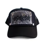 Gorra tipo trucker de color negro con panel frontal que presenta un parche rectangular con estampado de camuflaje y bordado estilo death metal, unido mediante costuras visibles en color blanco. Incluye un detalle de tachuela metálica piramidal en la base del panel frontal y parte trasera de malla.