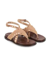 Sandalia plana de cuero gamuza color tostado, con tiras cruzadas estilo ojota adornadas con tachas metálicas doradas y tira ajustable al tobillo con hebilla.