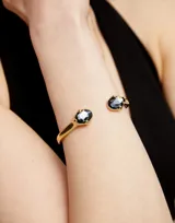 Pulsera rígida dorada de aleación de metales bañada en oro 18k, con dos cristales facetados grises en los extremos.