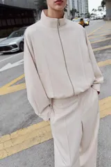 Cazadora estilo bomber de color beige, con cuello subido, manga larga y cierre frontal con cremallera. Presenta bolsillos delanteros de vivo y bajo ajustable con cordones elásticos laterales.