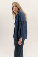 Camisa de jean azul oscuro con cuello mao, botones frontales, mangas largas abullonadas con puños elásticos y detalles bordados en color marrón en los hombros y mangas.