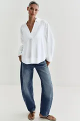 Blusa blanca confeccionada en lino, con cuello polo, escote en V y mangas largas amplias. Presenta un diseño holgado y bajo asimétrico.