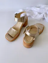 Sandalias de cuero color dorado, con tira ancha sobre los dedos, pulsera al tobillo con hebilla metálica y taco de madera.