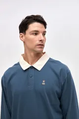 Remera polo de manga larga en color azul, confeccionada en algodón pesado con textura suave. Presenta cuello camisero en color crema, cierre frontal con botones y puños en las mangas. Incluye un pequeño bordado en el pecho.