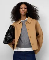 Chaqueta corta de color camel confeccionada en mezcla de lana y poliéster. Presenta cuello de solapa, manga larga de estilo japonés, cierre frontal con botones a tono y bolsillos laterales. Incluye un detalle de capa superpuesta en la espalda.