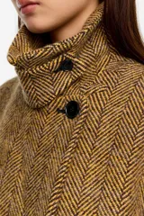 Abrigo largo de corte recto con diseño de espiga en tonos mostaza y negro. Presenta cierre frontal de doble botonadura, cuello con solapas clásicas y detalle de tira de ajuste en el cuello.