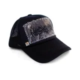 Gorra tipo trucker de color negro con panel frontal que presenta un parche rectangular con estampado de camuflaje y bordado estilo death metal, unido mediante costuras visibles en color blanco. Incluye un detalle de tachuela metálica piramidal en la base del panel frontal y parte trasera de malla.