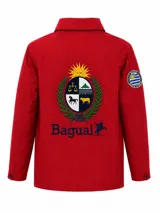 Campera impermeable de color rojo con cuello alto, confeccionada en tejido taslon resistente al viento y la lluvia. Presenta un gran escudo bordado en la espalda con motivos patrios y el logo de la marca Bagual, además de un parche circular en la manga con la bandera de Uruguay.