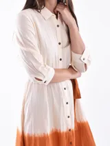 Vestido camisero midi de lino con diseño bicolor, presentando una parte superior en color crudo y una parte inferior en color naranja con efecto degradado. Posee cuello camisero, cierre frontal con botones y un lazo ajustable en la cintura del mismo tono que la parte inferior.