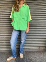 Camisa verde manzana de hilo de algodón, corte holgado, con cuello clásico, cierre frontal con botones, mangas cortas con doble raya amarilla y bolsillos de parche en el pecho.