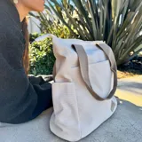 Cartera tipo tote de lona color marrón con asas de mano en color beige. Cuenta con un bolsillo frontal, bolsillos laterales y un detalle de tirador de cierre en cordón blanco.