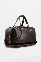 Bolso de fin de semana tipo duffle, confeccionado en material sintético efecto charol color marrón oscuro. Cuenta con doble asa superior, correa ajustable extraíble, cierre principal y logo de la marca estampado en el frente.