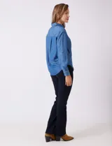 Camisa de tencel azul, marca Weekend Routine, con cuello solapa, botones metálicos frontales y manga larga regulable con presilla y botón. Presenta canesú horizontal en la espalda con tablón central y bajo asimétrico redondeado.