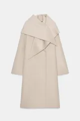 Abrigo de mezcla de lana color beige claro, de corte oversize y largo medio. Presenta un cuello solapa envolvente con detalle de pañuelo integrado y manga larga. Cierre frontal cruzado con botones ocultos.