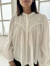 Camisa blanca de algodón con cuello mao, mangas largas con puños elásticos y cierre frontal de botones. Presenta un diseño con pliegues en la parte delantera y detalles bordados en el pecho.