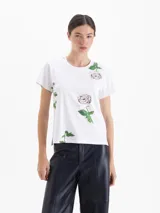 Remera de algodón blanca con estampado de rosas, cuello redondo, mangas cortas y detalle de tachas metálicas en los hombros.