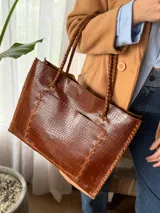 Cartera de cuero genuino color chocolate, tipo tote bag, con asas trenzadas, costuras artesanales visibles en los bordes y un amplio bolsillo frontal.