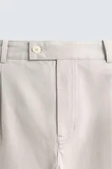 Pantalón de corte relajado confeccionado en tejido de algodón con estructura twill. Presenta pliegues frontales, bolsillos laterales y bolsillos traseros de doble vivo con botón. Cierre frontal con cremallera y botón.