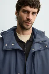 Parka de tejido técnico con acabado engomado, cuello subido y capucha ajustable. Presenta cierre frontal con cremallera oculta bajo solapa con botones a presión, bolsillos laterales de vivo y mangas largas con puños ajustables.