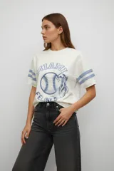 Remera de algodón color blanco con estampado frontal estilo deportivo en tonos azules, cuello redondo y mangas cortas con detalle de rayas horizontales.