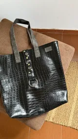 Tote bag de gran tamaño confeccionada en ecocuero negro con textura de reptil. Presenta una tira central decorativa con ojales metálicos y una argolla grande en la parte inferior, además de manijas en color gris.