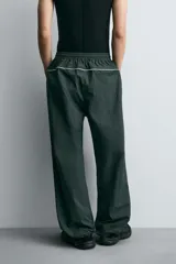Pantalón jogger de corte relajado confeccionado en tejido técnico de color verde oscuro. Presenta cintura elástica con cordón interior, bolsillos laterales con cierre y bajo ajustable con elástico. Incluye detalles de líneas en contraste a lo largo de las piernas.