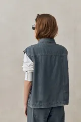 Chaleco de jean azul con sutil textura a rayas, cuello camisero, cierre frontal con botones metálicos y diseño sin mangas con hombros amplios.