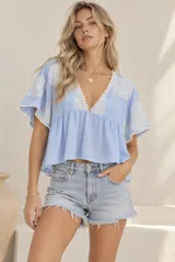 Blusa celeste con escote en V, mangas cortas amplias y diseño fluido. Cuenta con detalles de bordados florales en blanco en el escote y las mangas, además de terminaciones con pequeños pompones.