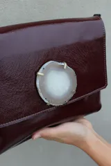Sobre de mano color bordó en cuero charolado, con solapa frontal decorada con una piedra ágata natural engarzada. Incluye correa larga extraíble y cierre trasero.