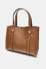 Bolso tipo mini shopper de cuero sintético color marrón caramelo, con costuras blancas visibles. Posee asas de mano y una correa bandolera ancha de tela color beige. El frente tiene un detalle de tira con hebilla metálica dorada.