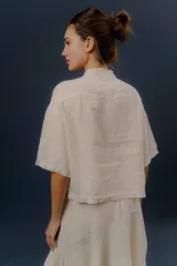 Blusa de lino color crudo, con cuello mao, mangas cortas y cierre frontal cruzado con nudos de tela y botones.