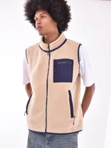 Chaleco de polar tipo corderito en color beige, con cuello alto y cierre frontal completo. Presenta un bolsillo de parche en el pecho en color azul marino con cierre de cremallera y ribetes en contraste en el mismo tono en sisas, cuello y bolsillos laterales.