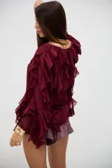 Blusa bordó de tela semitransparente con diseño de volados verticales en el frente y mangas largas acampanadas. Presenta cierre frontal con botones y detalles de bordados circulares sutiles en la parte inferior.