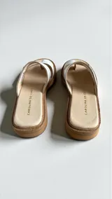 Sandalias de cuero blanco modelo Lena de Carolina De Cunto, con diseño de doble tira, sujeta dedo y detalle de aplique metálico y pintura artesanal multicolor en la tira frontal. Cuenta con base acolchada y suela con textura de yute.