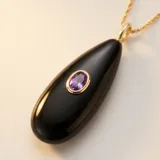 Collar de acero con baño de oro, compuesto por una cadena fina y un dije en forma de gota de piedra natural ónix negra con una circonia violeta engastada en el centro.