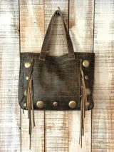 Cartera estilo shopper de cuero con textura de cocodrilo, color marrón oscuro. Presenta detalles de tachas metálicas circulares y flecos laterales de gamuza. Cuenta con doble asa superior.