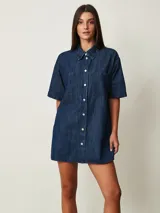 Vestido corto de denim azul oscuro, con cuello camisero, mangas cortas, cierre frontal de botones y dos bolsillos en el pecho.