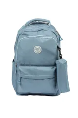 Mochila escolar de tela color azul claro, con múltiples compartimentos con cierre, bolsillo frontal con cierre y un pequeño accesorio colgante cilíndrico a juego. Presenta un parche circular con un emoji sonriente en el frente.