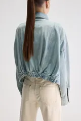 Sobrecamisa oversize de denim celeste con acabado lavado, cuello clásico, cierre frontal con broches metálicos, bolsillos frontales con solapa, mangas largas con puños abotonados y detalle elastizado en el ruedo posterior.
