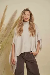 Poncho tejido de punto acanalado en color beige, con cuello alto, diseño irregular y terminación con flecos en los bordes.
