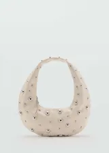 Bolso de hombro con diseño ovalado tipo hobo, confeccionado en material efecto piel color beige, decorado con tachuelas metálicas plateadas distribuidas por toda la superficie y cierre de cremallera.